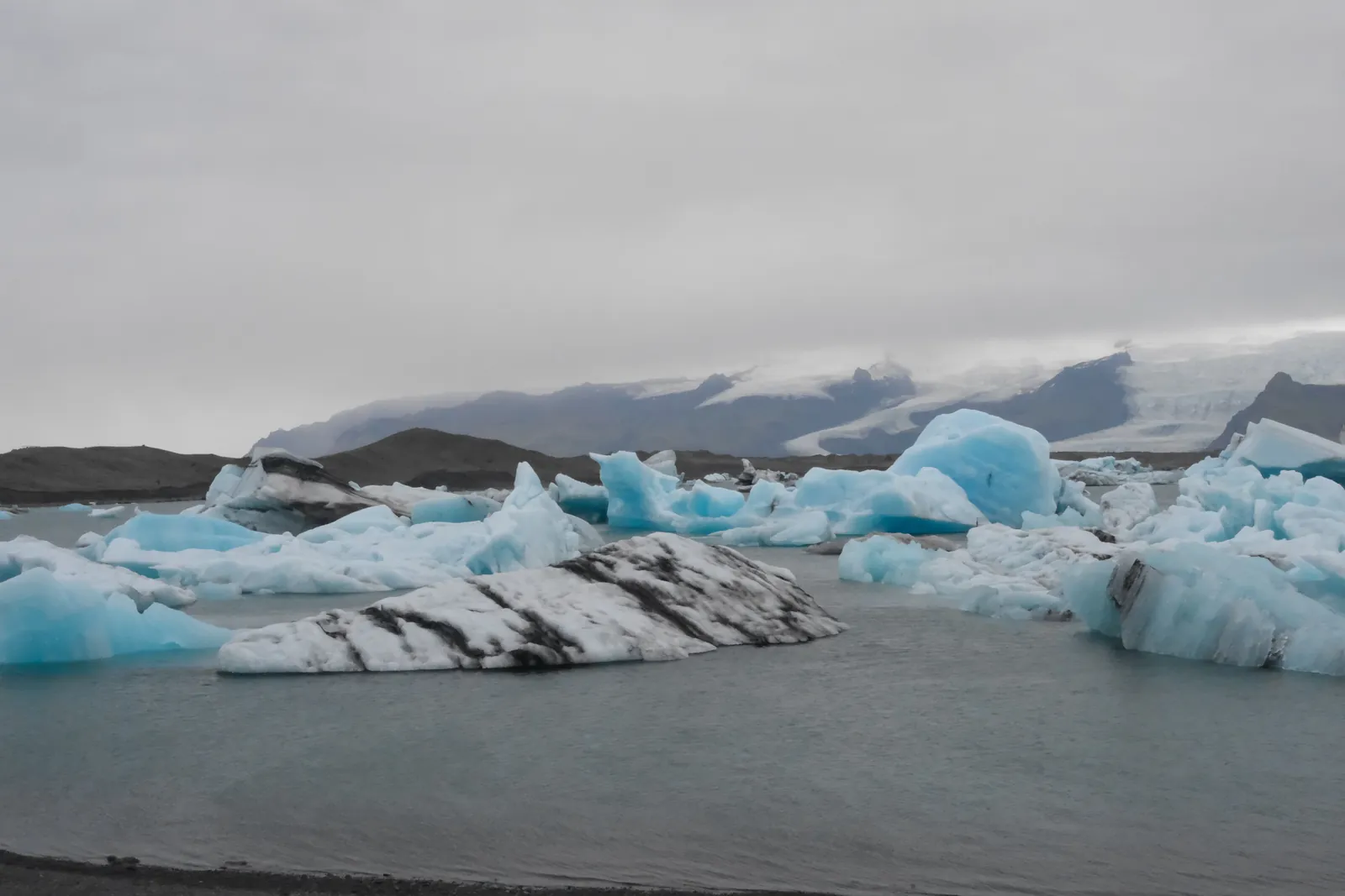 Blue icebergs float in the calm waters of Jökulsárlón Glacier Lagoon, Iceland, with distant mountains under a cloudy sky.