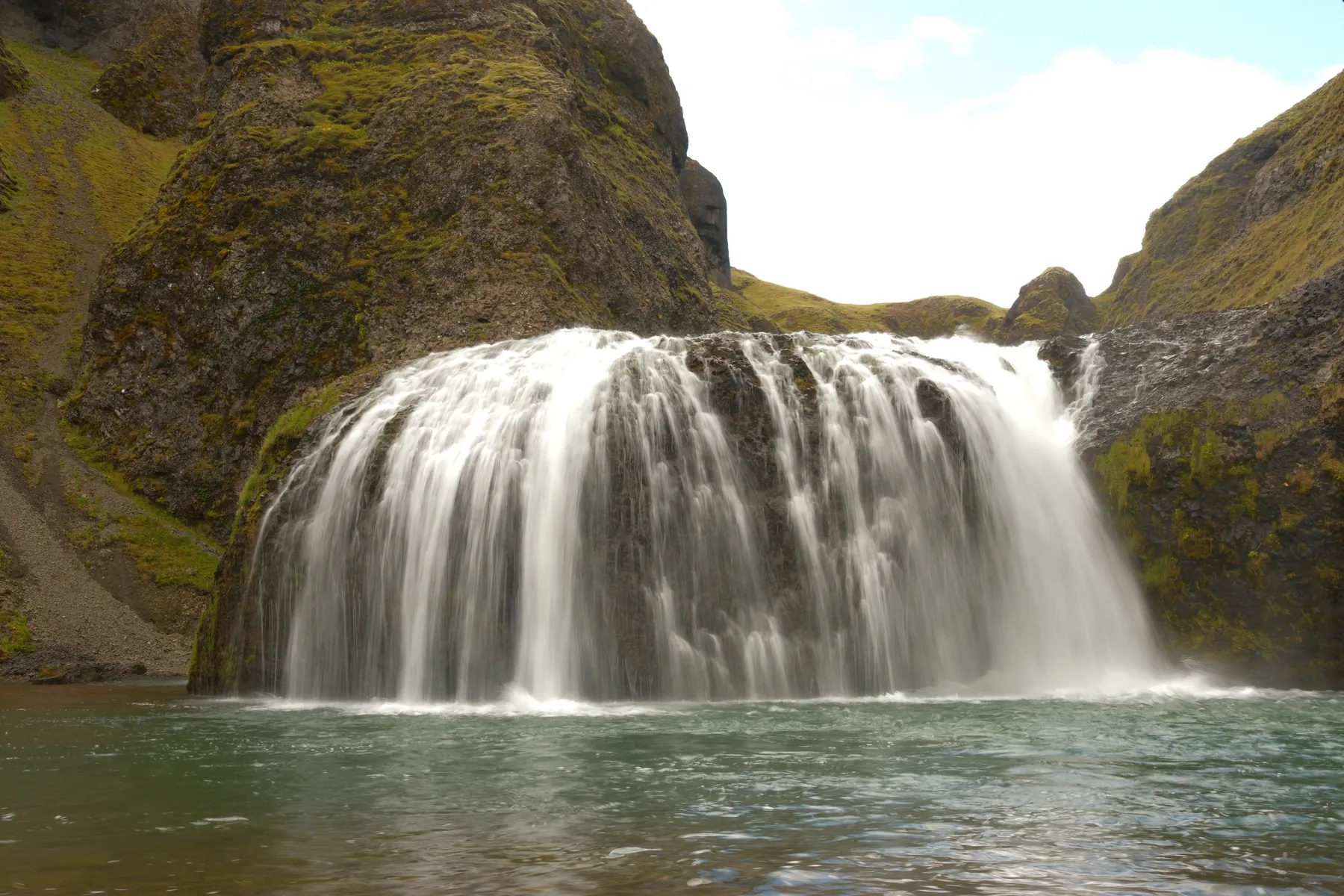 A wide waterfall flows over a rocky ledge into a turquoise pool.