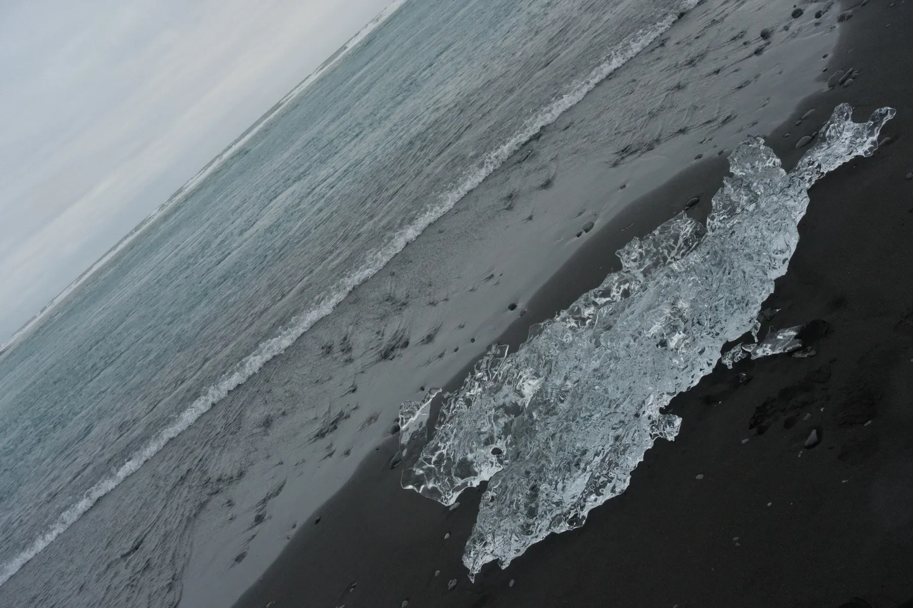A clear ice fragment rests on a black sand beach with gentle waves.