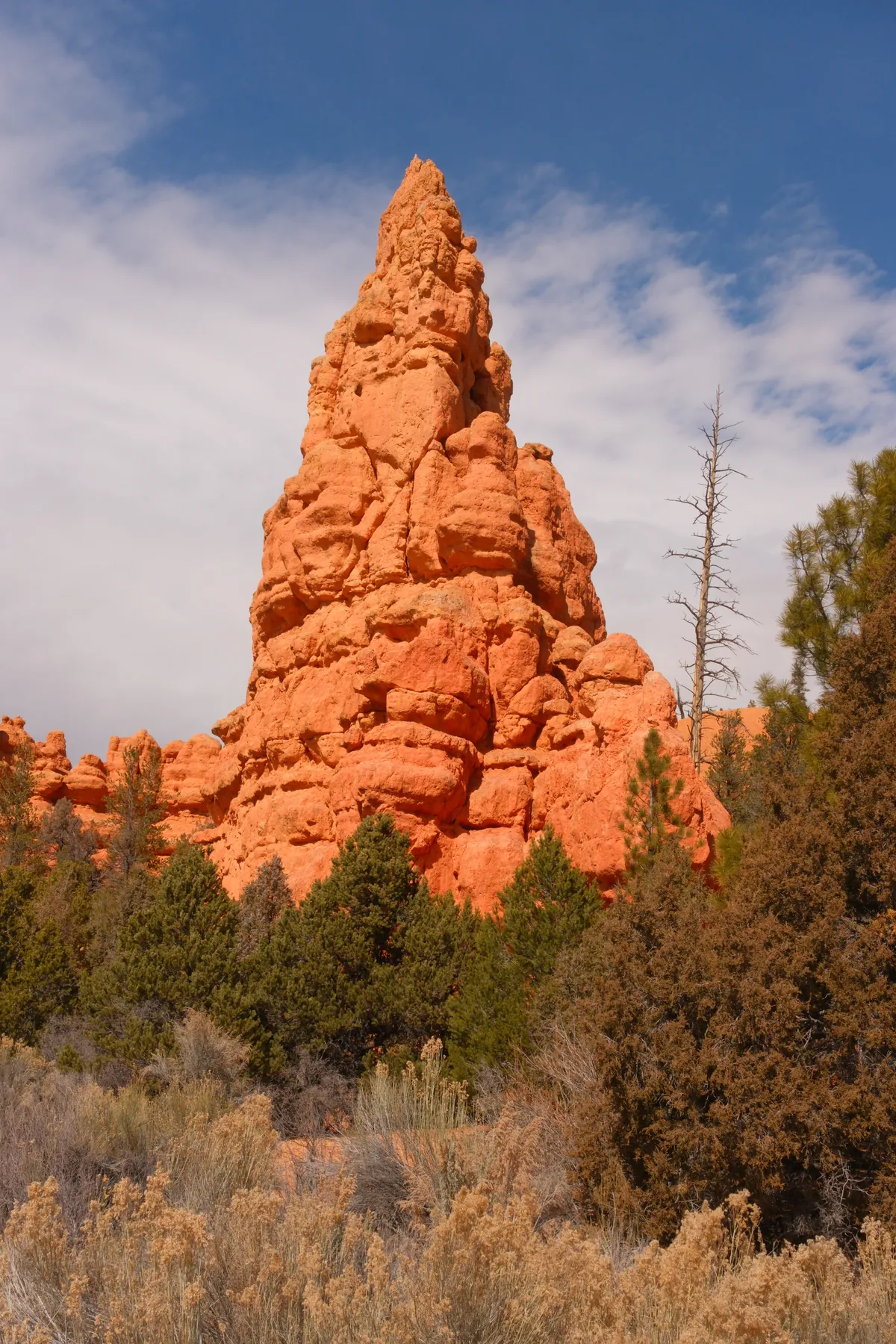 A tall, eroded red rock spire stands prominently against a blue sky with scattered clouds.