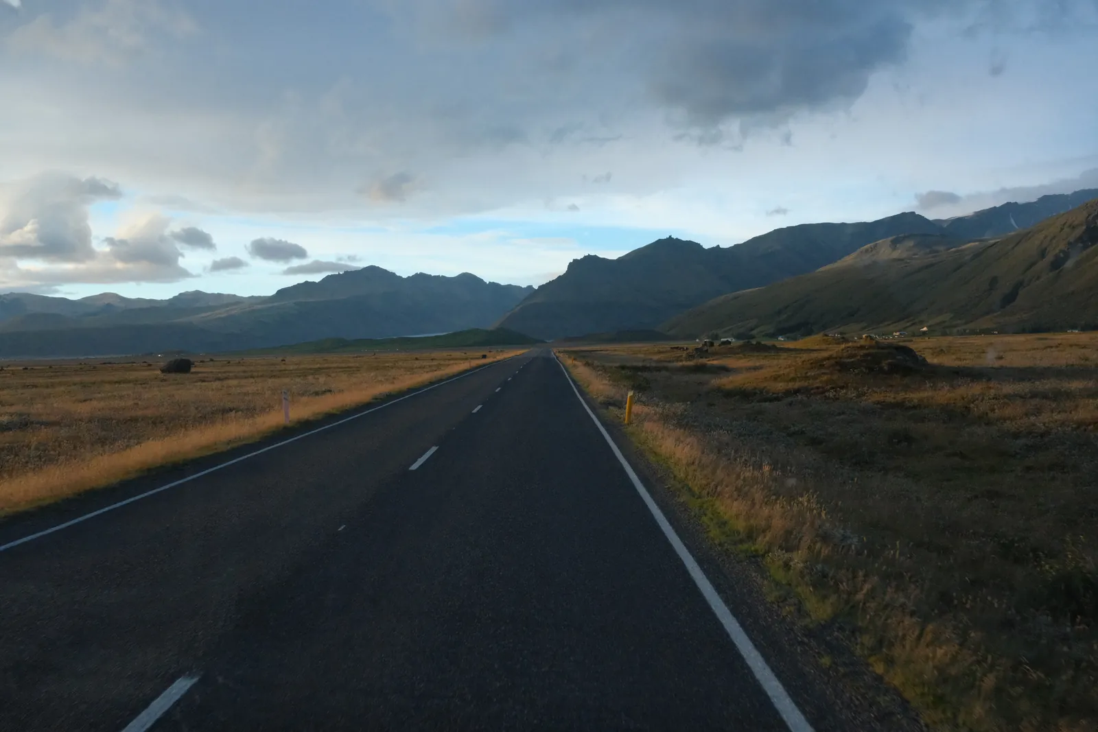 A paved road stretches through a vast, grassy plain towards distant mountains under a cloudy sky.