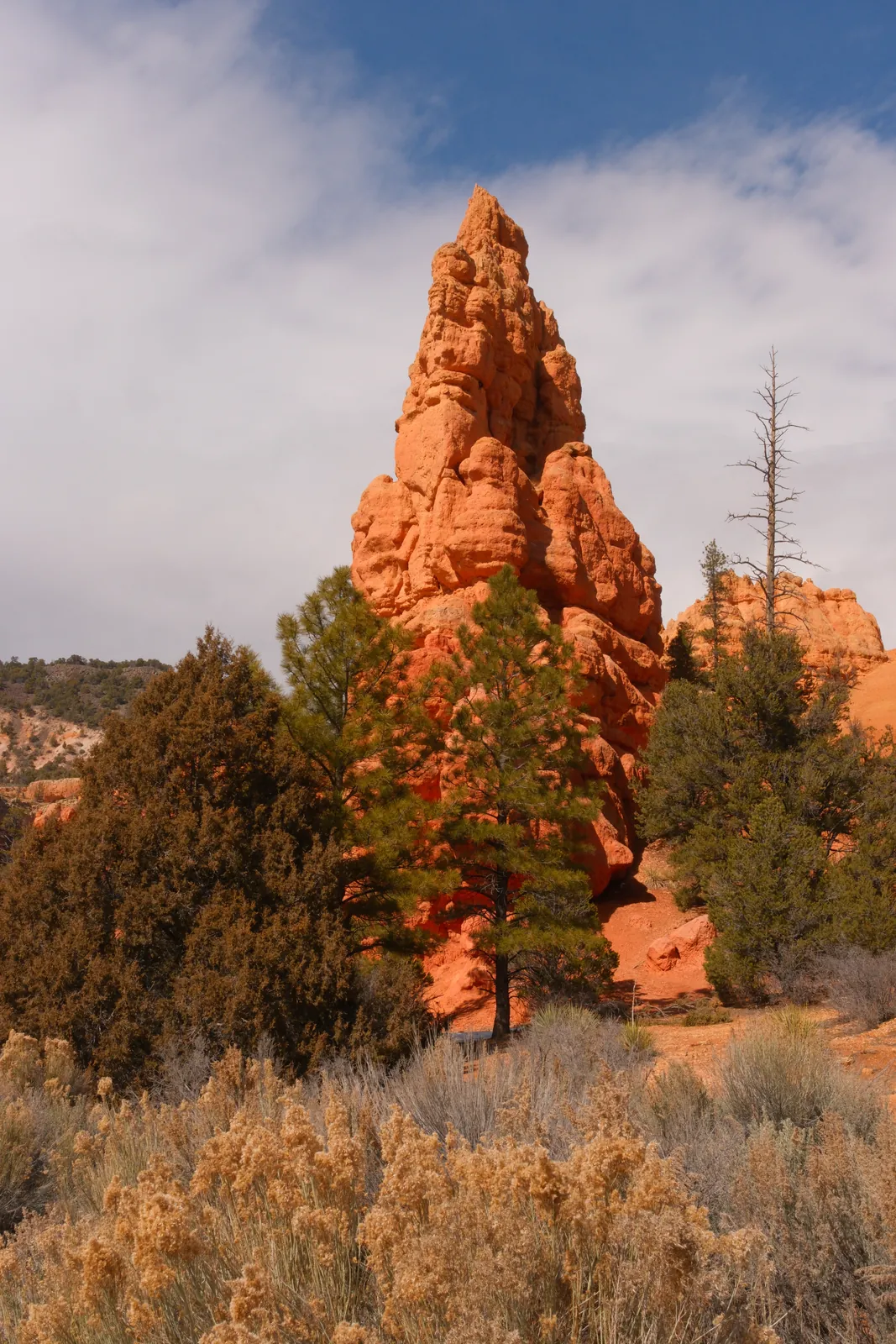 A tall, orange rock spire stands prominently against a cloudy blue sky.
